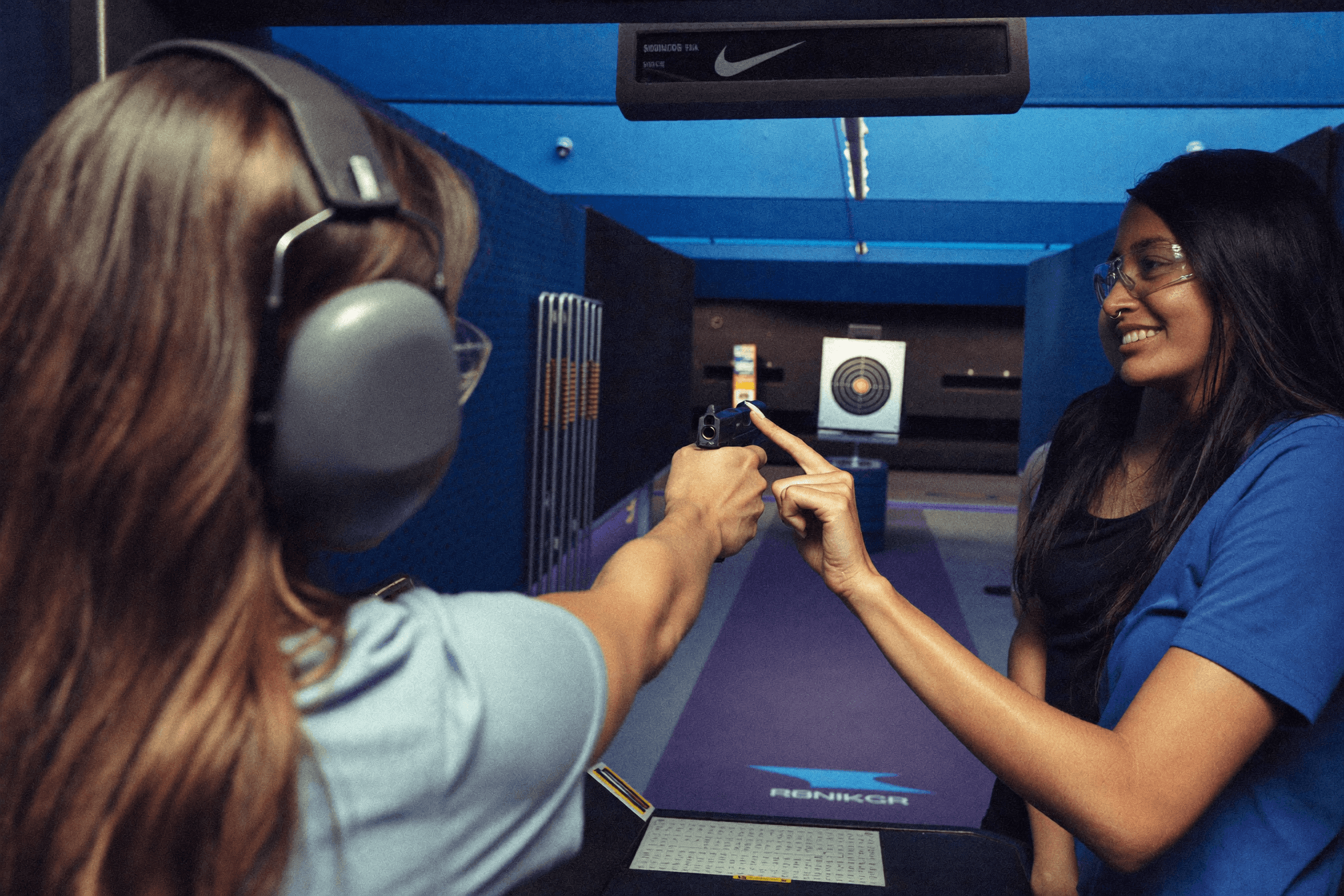A first-timer woman aiming down an indoor shooting range lane with a friend behind her, target downrange, cool blue tone.