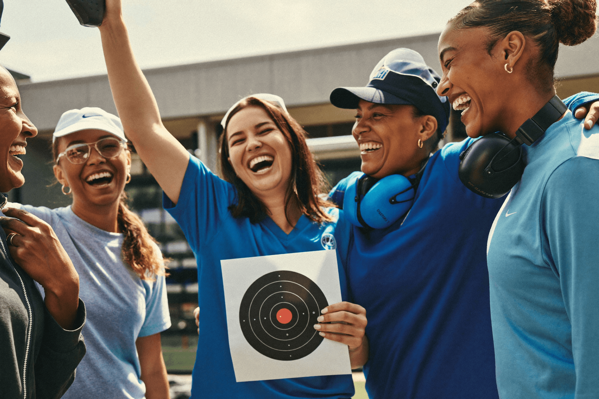 A group of women shooters laughing together at a range, one holding up a target, in a cool blue tone.