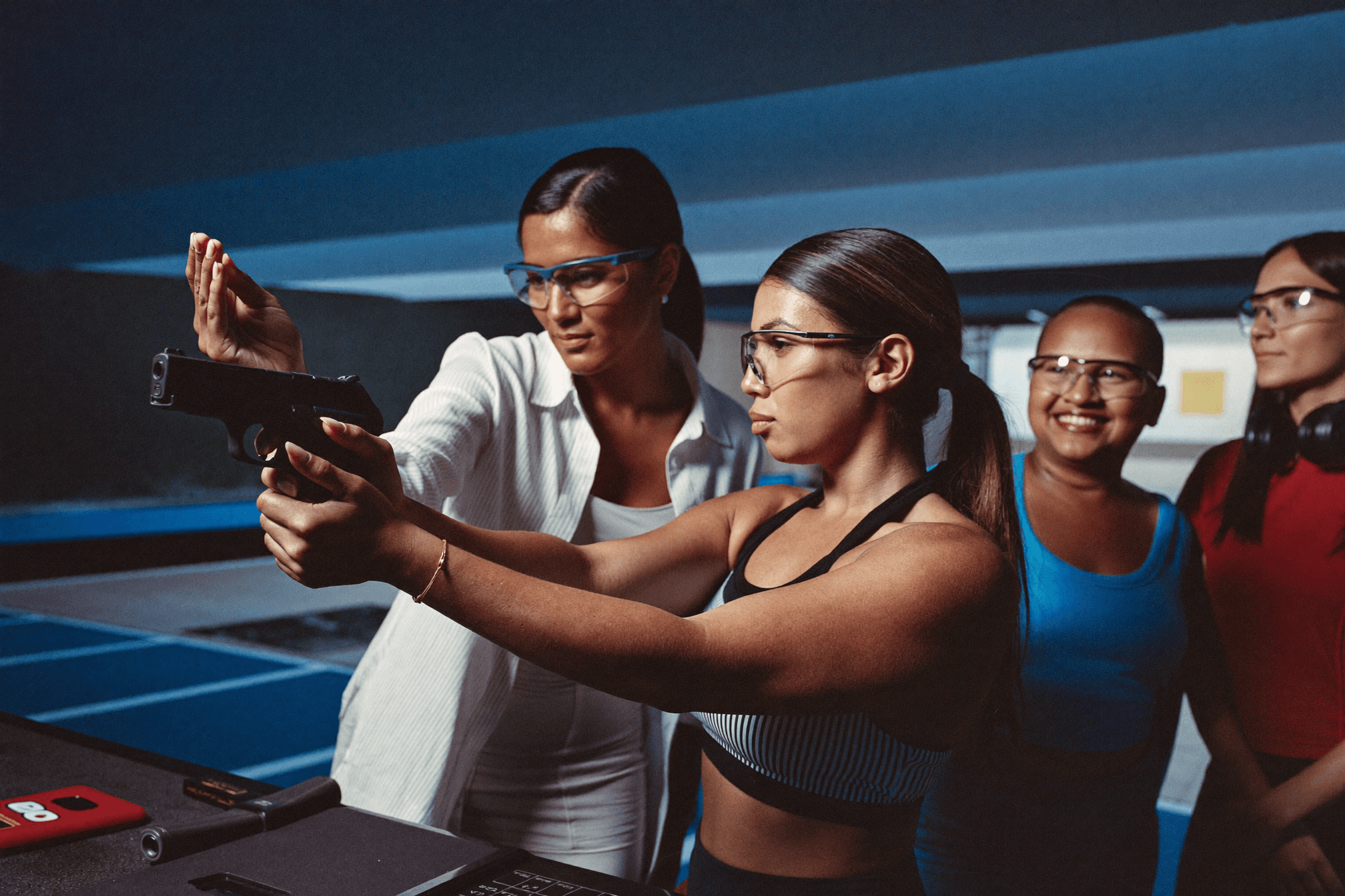 Female instructor coaching women on the firing line at a shooting range, all in eye and ear protection, in a cool blue tone.